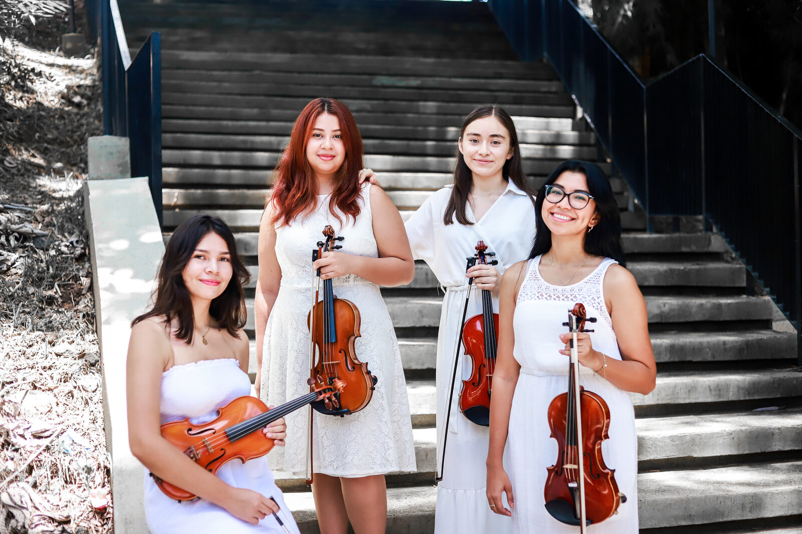 Cuatro violinistas de la Sociedad de Música de Cámara de El Salvador, vestidas de blanco, en una escalinata al aire libre